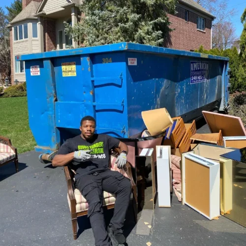 Man sits by a large blue dumpster with furniture, Rubble Removers logo visible. Rubble Rumblers