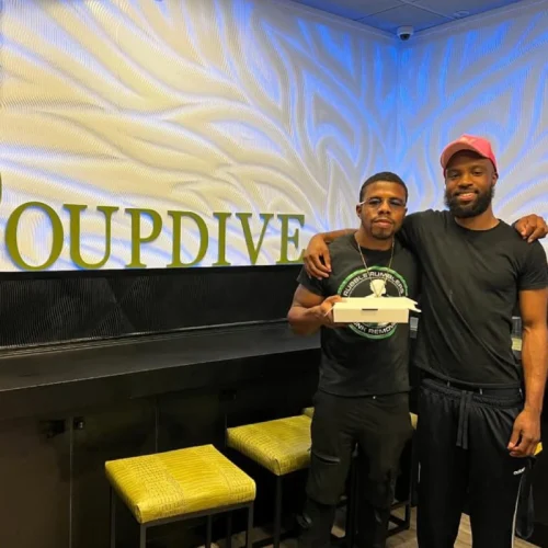 Two men pose in front of a wall with "GROUPDIVE" text, one holding a takeaway container. Rubble Rumblers
